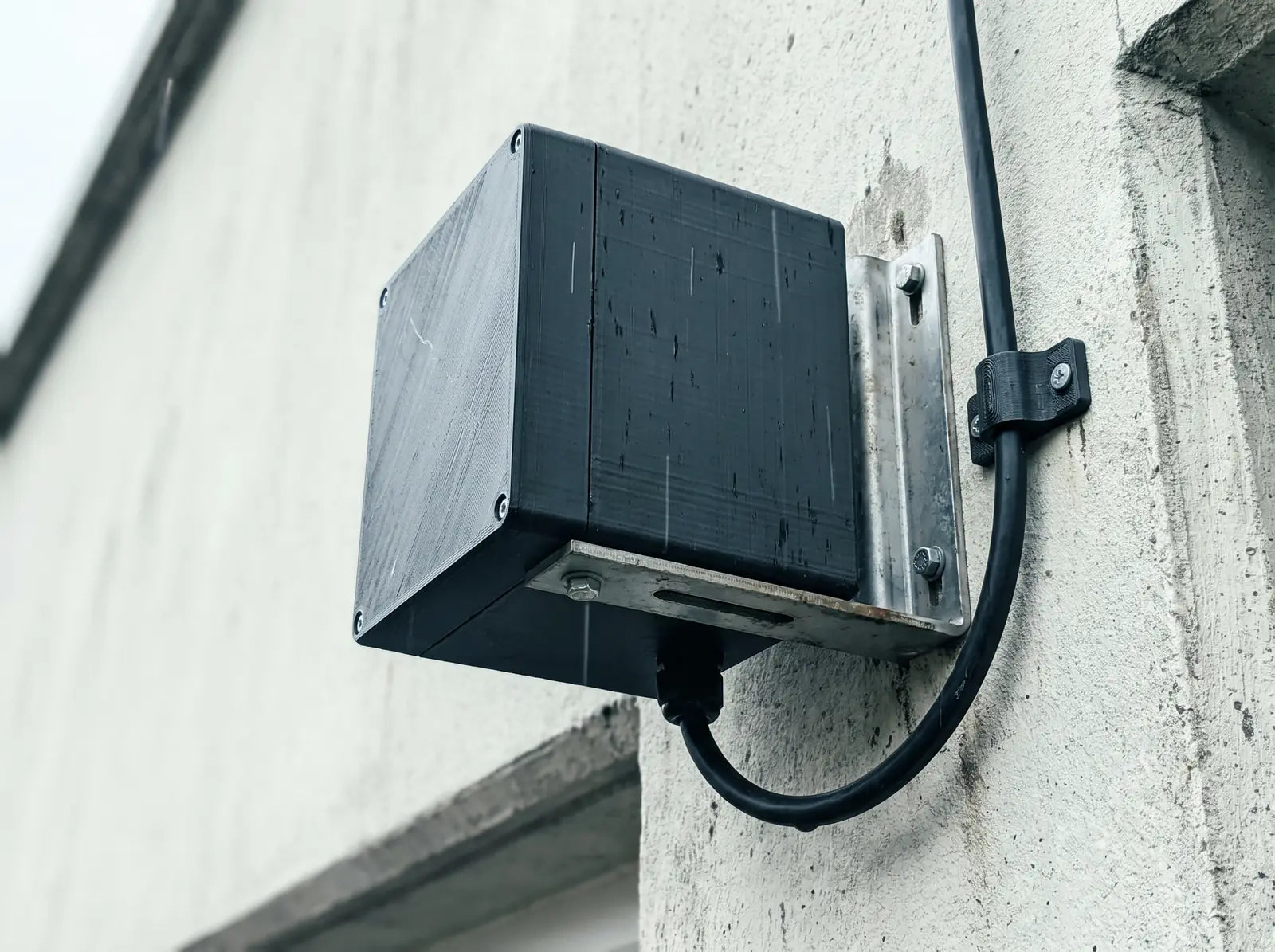 A 3D-printed enclosure on a steel wall bracket outdoors, weather-exposed, with a printed cable clip holding the cable run beside it
