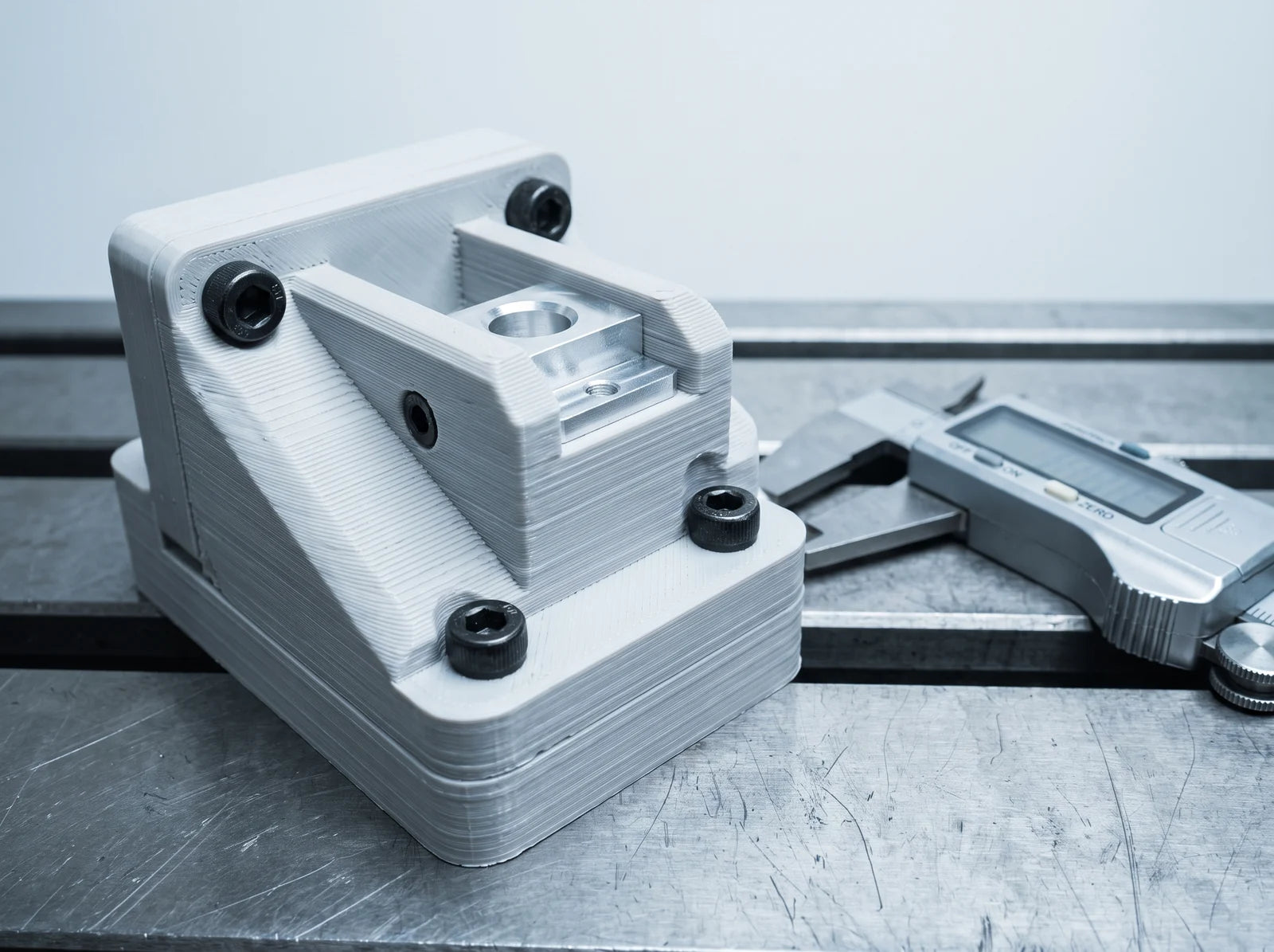 A 3D-printed fixture bolted to a machinist's steel bench, holding a machined aluminium part in position, with digital calipers alongside